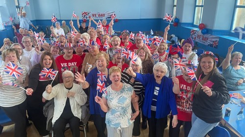 A large group of people in a community centre hall cheering and celebrating, waving small Union Jack flags