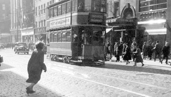 Black and White photo of a street with a tram and a older women crossing the road, in the background there is a car and a crowd of people. The entrance to the Argyle Arcade can be seen to the right of the tram.