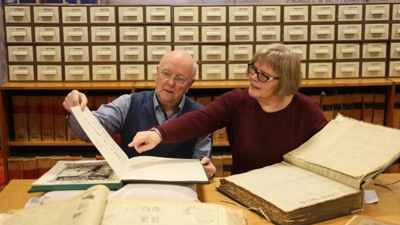 Two people sitting at a desk with two large old books in front of them with filing cabinets behind them in the background