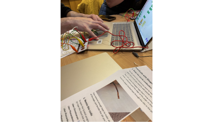 A person typing on a laptop with an attached microcontroller circuit board and alligator clips, while an instruction sheet titled "ChroniCode with Glasgow" lies in the foreground.