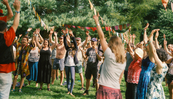 A large group of people participate in an outdoor laughter yoga session on a green lawn, raising their arms together in a synchronised gesture.