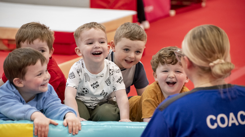 A group of children are smiling and laughing while leaning on gymnastics equipment. A person in a blue coach top is at the front of the photo.
