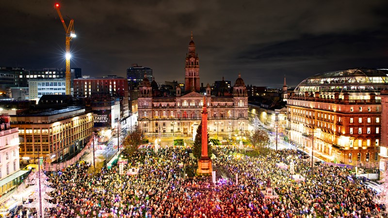 An aerial shot shows thousands of people gathered in a large civic square with a large monument in the middle and festive lighting round the perimeter