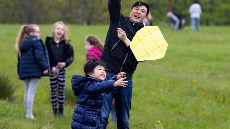 A child and adult laugh together as they play with a kite. Behind them, in a grassy setting, more children laugh together