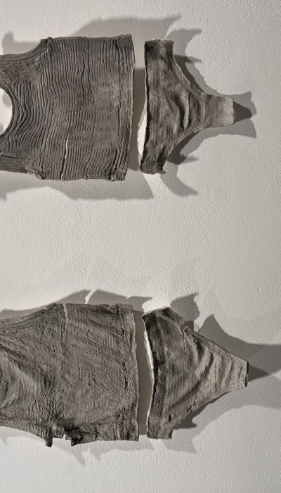 Two sculptural pieces resembling the upper and lower halves of sleeveless tops are mounted on a white wall. Each form is made from a grey, textured clay. The upper piece has a ribbed, fabric‑like pattern, while the lower one has a smoother, more organic surface. Both are horizontally split, creating a clear separation between the top and bottom sections. Soft lighting casts defined shadows that emphasise their three‑dimensional shape and surface detail.