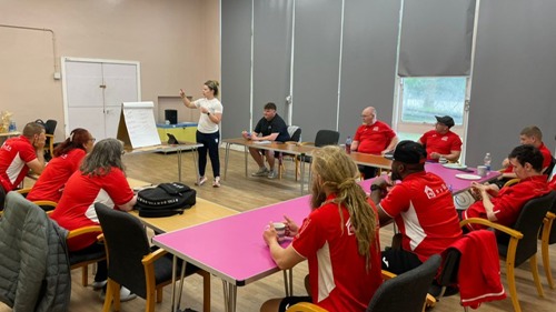 A group of people in the same red tshirts taking part in a training course at a community centre. They are all sat a desks, looking at an instructor at a flipchart