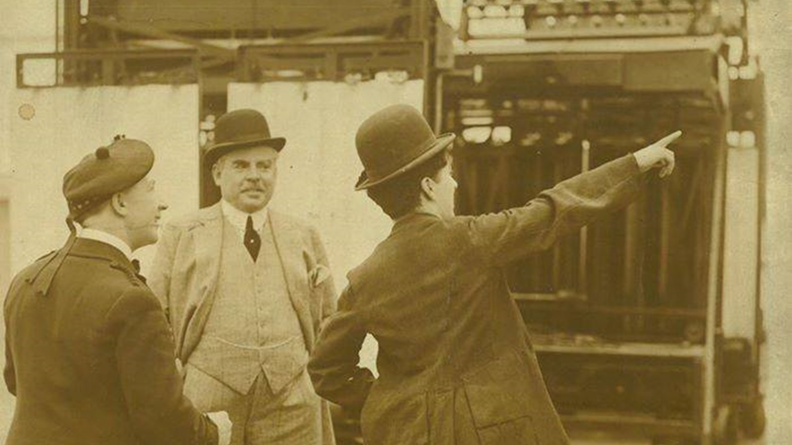 A black and white photograph of three people, including Harry Lauder and Charlie Chaplin in a warehouse, all wearing hats and one pointing to something in the distance.