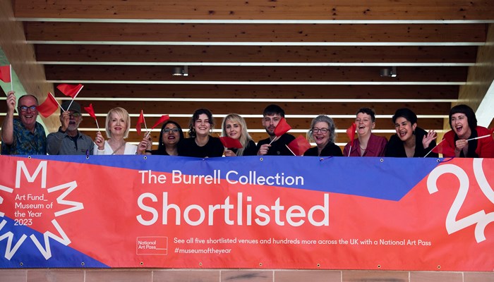 Staff at The Burrell Collection stand waving flags with a giant banner celebrating being shortlisted for Art Fund Museum of the Year 2023