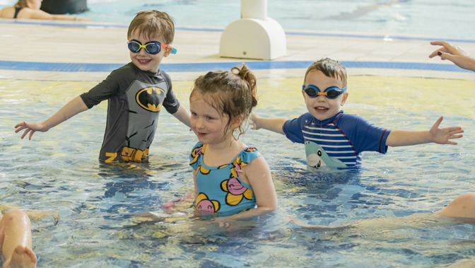 Three young children playing in a swimming pool, two are wearing goggles and have outstretched arms.