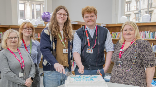 A group of five people gathered around a large white cake to celebrate the 100 anniversary of the library they are standing in