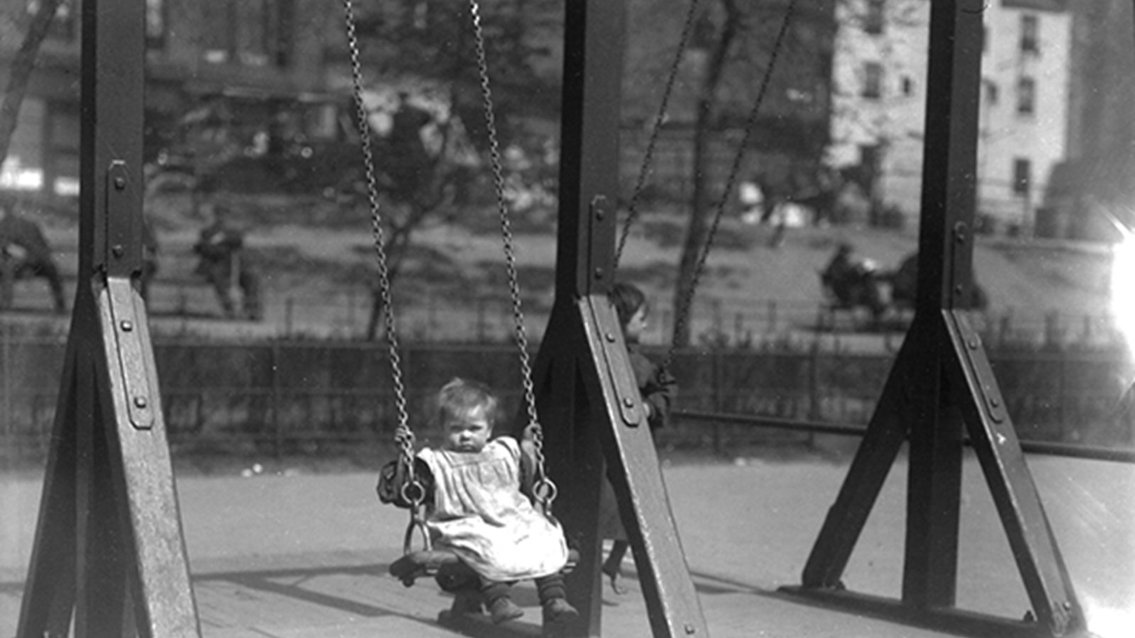 A black and white photograph of a baby on a swing holding onto the swing chain, wearing dirty clothes. There is an older child on the other swing and buildings in the background.