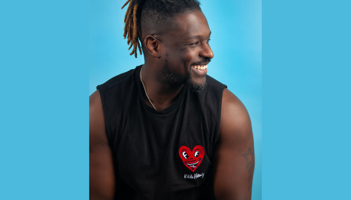 A smiling man with short hair and dreadlocks pulled back looks to his right, wearing a black sleeveless top featuring a Keith Haring heart graphic, set against a bright blue background.