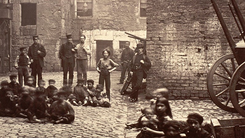 Black and white image of a Glasgow slums from 1800s, children are sitting on the ground, poorly dressed and adults stand in the background