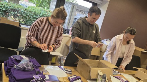 Three people are working together behind a table to put promotional material together into bags for a charity event
