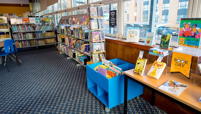 A corner of the children's area near large windows. There are bookshelves filled with colourful books, book displays and a poster on the window that reads 'Free WiFi'