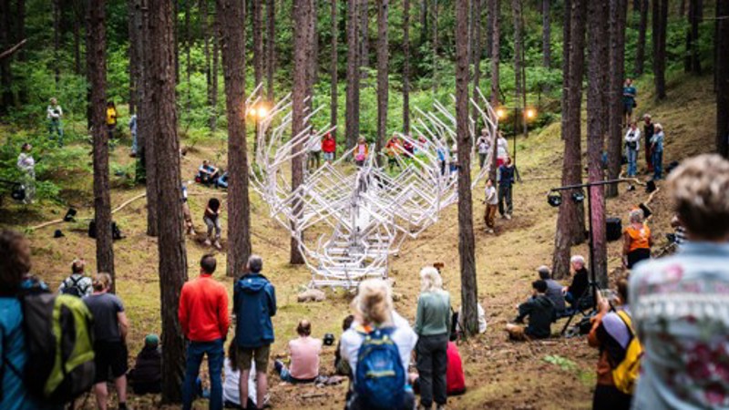 Visitors stand among tall pine trees watching a large white sculptural installation in a woodland clearing.