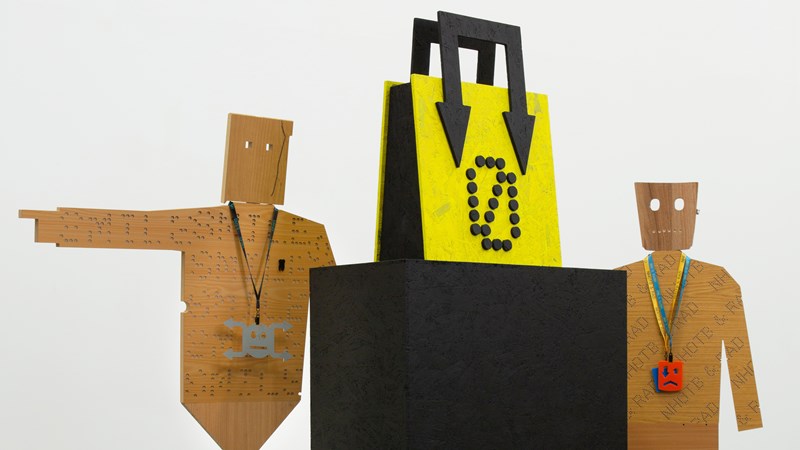 Wooden cut-outs are placed either side of a large yellow handbag placed on top of a black box