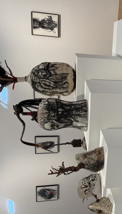 A gallery display features a group of mixed‑media sculptures arranged on white pedestal platforms. The pieces vary in size and shape, resembling abstract vessels or organic forms made from materials such as clay, wood, fur, and painted surfaces. Several sculptures have protruding sticks, feathers or branches. Behind the sculptures, framed artworks hang on the wall.