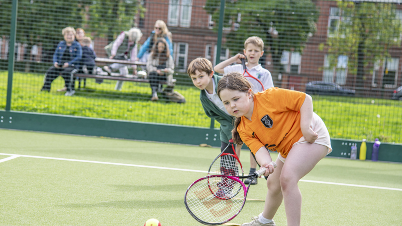 A child in an orange tee-shirt hits a tennis ball with a racket. A queue of children await their turn to hit the ball.