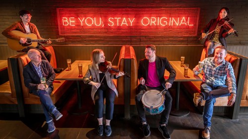 A group of six people are gathered around two booths in a pub. Two of them are holding violins, one a guitar and one a drum. There are glasses of beer in front of them and a red neon sign on the wall behind that reads Be You, Stay Original