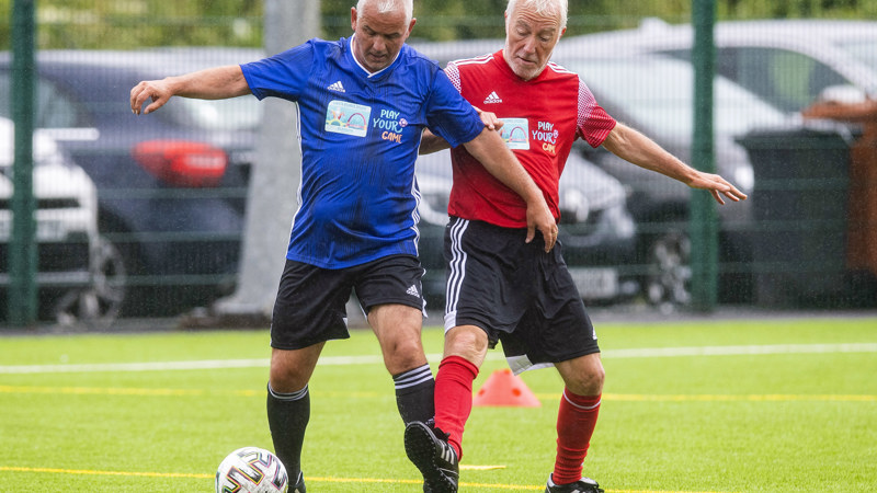 two men tackling each other on an outdoor football pitch with a carpark in the background, one man has the ball at his feet