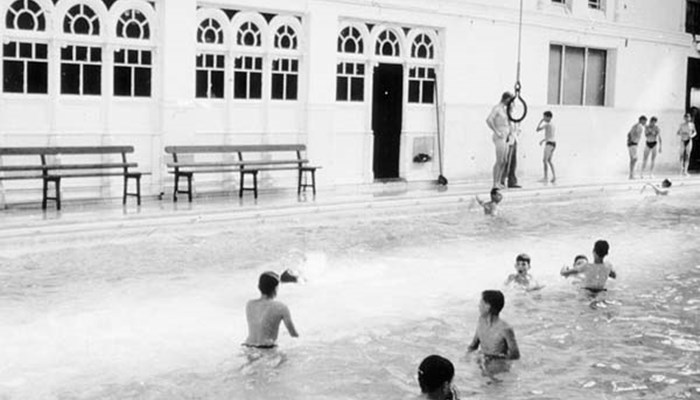 A black and white photo of people enjoying a swim in a rectangular swimming pool, with benches and gymnastic hoops in the background.
