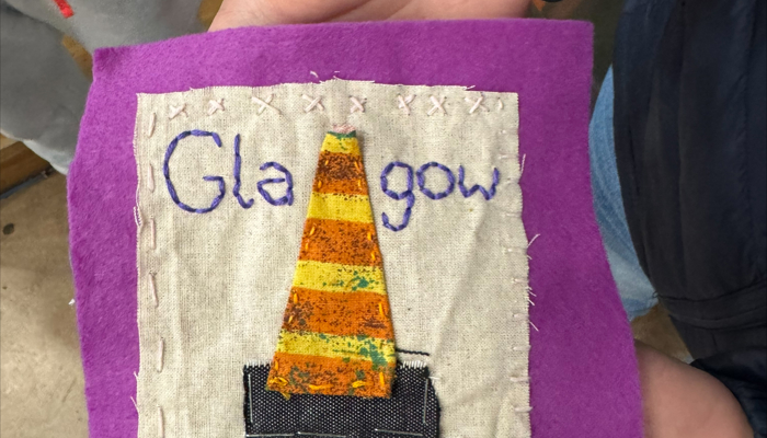 A small handmade textile piece held in one hand. A beige square of fabric is stitched onto a larger purple felt square. The beige fabric has the word “Glasgow” embroidered in blue at the top, with a fabric traffic cone below it made from orange‑and‑yellow striped material with a black base.