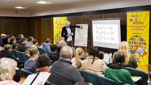 A person wearing a bow tie is drawing on a flipchart in front of an audience of seated adults and young people in a library
