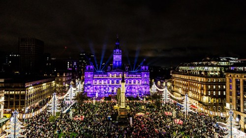 A large crowd is gathered in a city square to celebrate the Christmas lights switch-on. It is evening and the square is brightly lit with colourful decorative lights on all sides