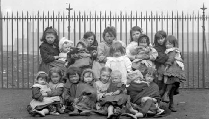 Black and white photo showing a large group of children and babies sitting together looking at the camera in a park with railings in the background, an aerial view of the city of Glasgow is also in the background