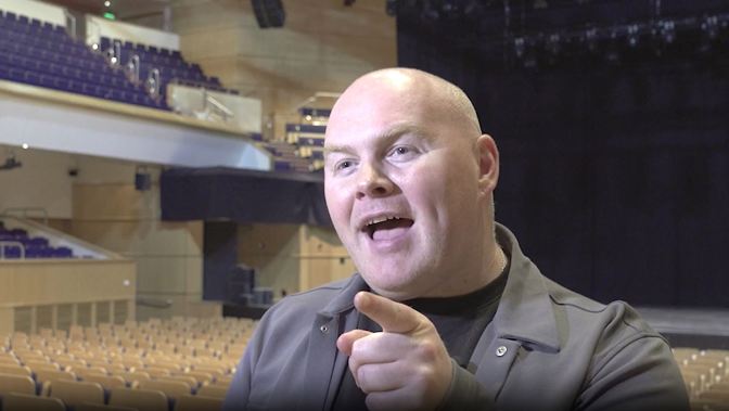 A bald man in a dark jacket smiles and points while seated in an empty theatre.