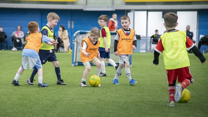 A group of young children are wearing team bibs on a football pitch, at the centre of the image a girl is about to kick the ball