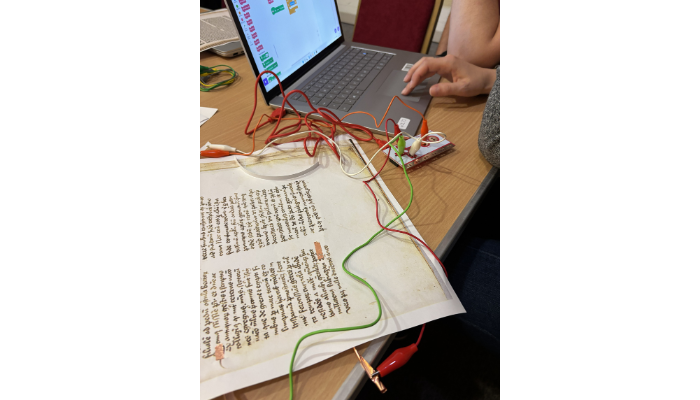 A person operating a laptop connected via colourful alligator clips and wires to an interactive circuit board and a printed sheet of manuscript text.
