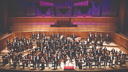A large orchestra standing with their instruments on a stage. They are all wearing black
