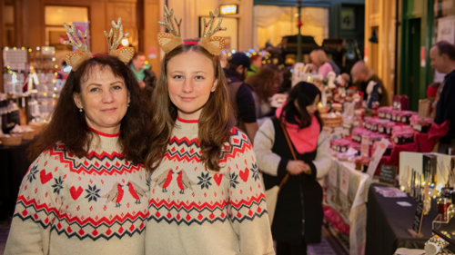 Two people wearing the same knitted Christmas jumpers and wearing reindeer antler headbands smiling in front of an indoor Christmas market