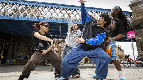 Four street dancers perform expressive, energetic moves beneath a blue iron bridge in an urban area. They wear casual, loose-fitting clothes and sneakers, striking dynamic poses that convey rhythm and creativity. Behind them, stone buildings and a pink sign reading “People Make Glasgow” highlight the lively city backdrop.