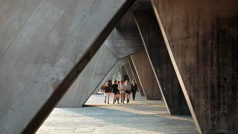 A group of children walk beneath a series of massive, angled concrete beams that form a dramatic covered walkway. The slanted supports create a geometric corridor of light and shadow across the paved ground. The scale of the structure towers over the children, emphasizing the bold, sculptural architecture surrounding them.