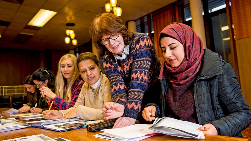 group of women of different nationalities sitting around a desk reading and writing with one women leaning over to check their work