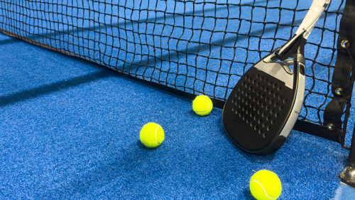 A padel racquet is propped up against a net on a padel court. Three padel balls are beside the racquet.