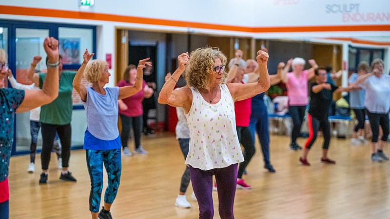 A group of ladies taking part in indoor fitness class all with their arms in the air.