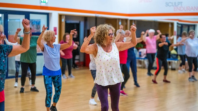A group of ladies taking part in indoor fitness class all with their arms in the air.