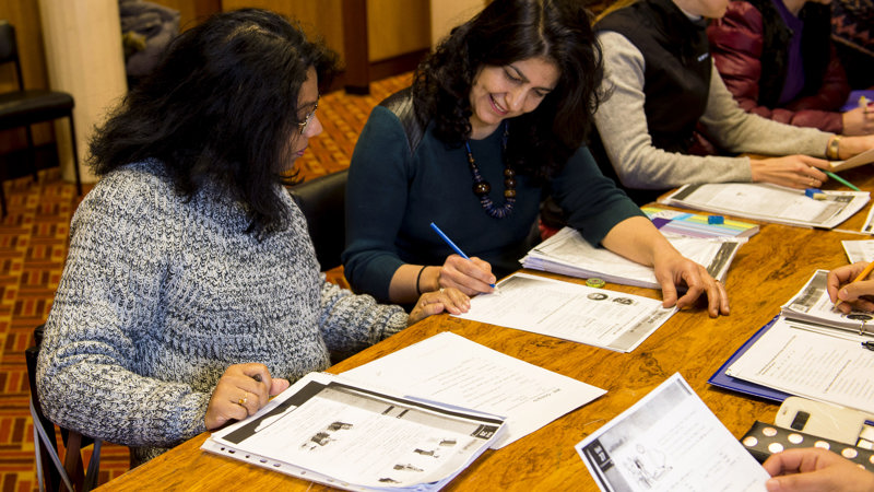 4 women from different ethnic backgrounds sitting at a desk with books, pens and paper in front of them