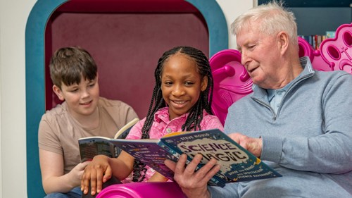 Two young people and an adult are sitting on the same large purple chair in a library. They are all looking at the same book and smiling