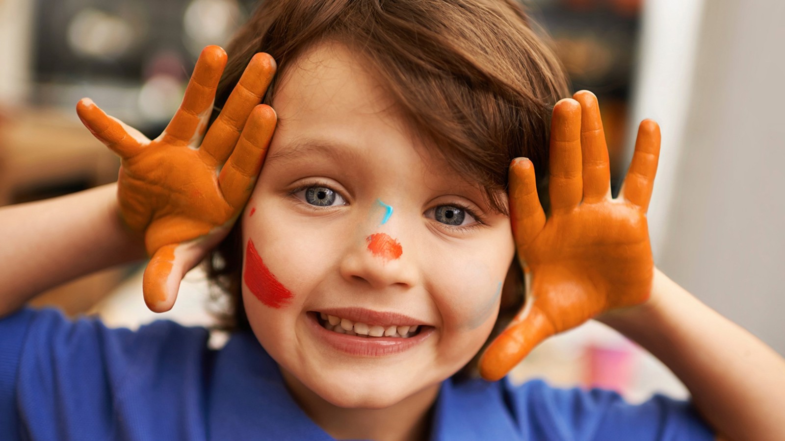 a young child looking into the camera. they have orange paint on their hands and face as they join an arts and crafts session