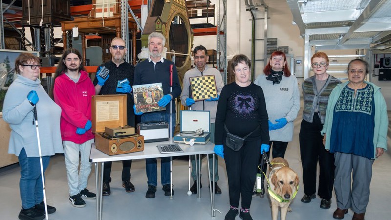 A group of people stand together indoors around a table displaying vintage items like a record player, a Beatles vinyl album, a chessboard, and cassette tapes. Some individuals hold white canes, and a guide dog stands in the foreground.