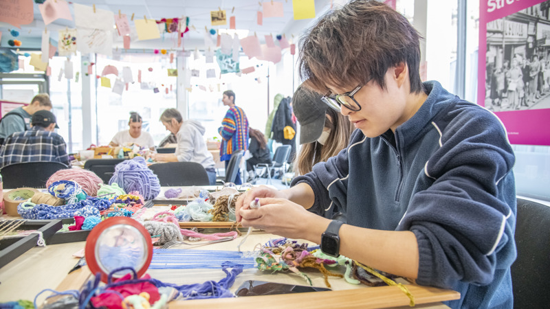 A picture of a person weaving on a loom in a busy and colourful room of people taking part in craft activities.