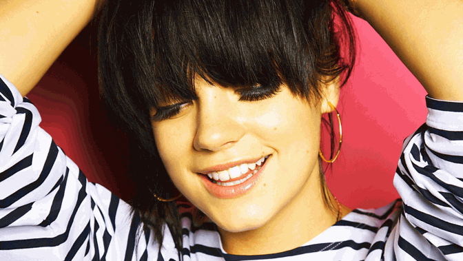 A close up of a persons face - they are smiling, looking down with their arms above their head and wearing a striped top