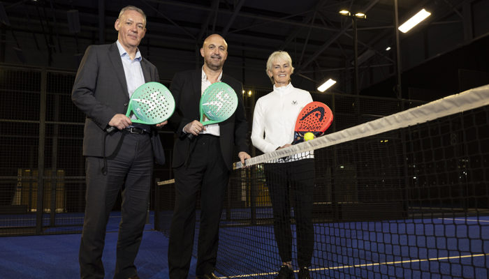 Two men and a woman stand either side of the net on a padel holding padel racquets, the woman is also holding a padel ball