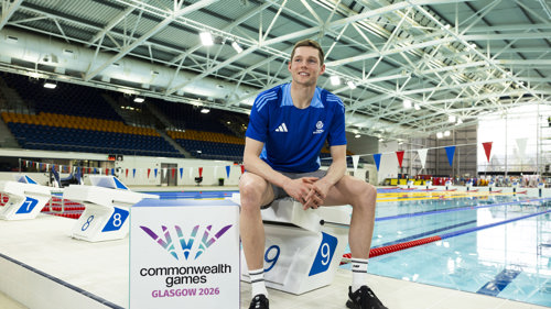 Swimmer Duncan Scott, wearing shorts and t-shirt is sitting on a starting block in an indoor swimming pool