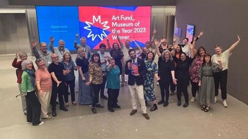 A large group of people celebrating with their arms in the air in a large museum space. One of them is holding a class trophy and there is a large screen behind them with text 'Art Fund Museum of the Year 2023 Winner'